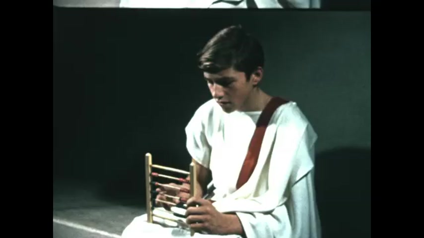 1950s: Boy in toga sits and manipulates abacus. Girl and woman in ancient style clothing sit and sew fabric.