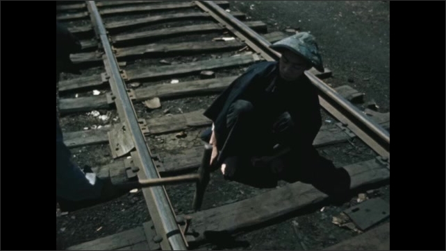 Decade: workers build railway line tracks with wood and metal nails. Man in hat crouches on ground. Man hits sleeper nail with mallet.