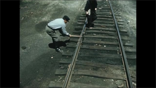 Decade: migrant workers build railroad track by hand. Man holds nail in sleeper. Man strikes nail with mallet. 