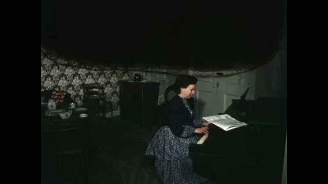 1850s: Woman plays piano. Man picks up newspaper and takes off jacket. Man sits.