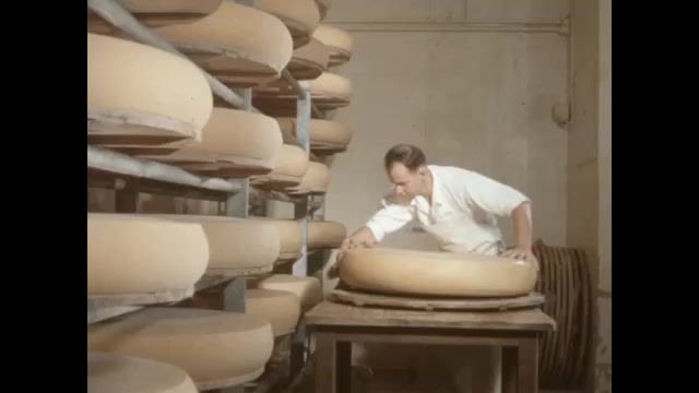 1950s: Cheesemaker wipes down large wheel of cheese and stacks it on shelf. Men unload smaller wheels of cheese in crowd.