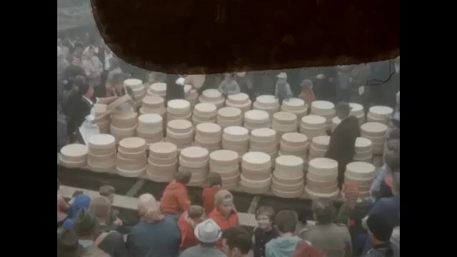 1950s: Men unload smaller wheels of cheese in crowd and stack them on tables. Alps above small village in valley.