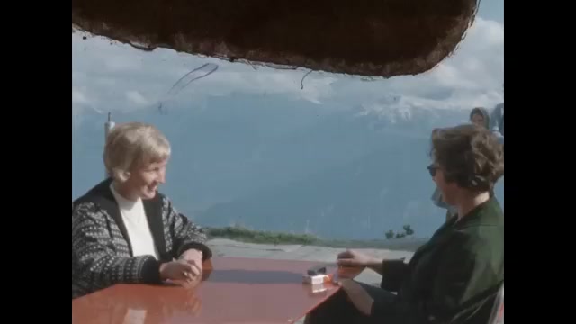 1950s: Two women are served coffee at outdoor café. People travel up and down mountains on chair lift. Snow capped mountains against sky.