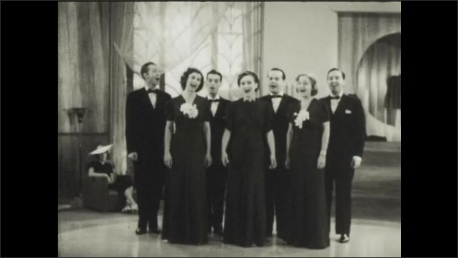 1930s: Group of three women and four men stand together on floor at performance, singing together. They finish song and bow. Man claps and walks onto floor.