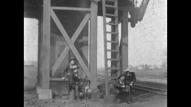 1900s: Robbers hide behind building.  Train stops.  Man uses chute to fill train car.