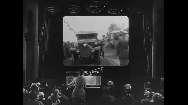 1910s: Movie theater.  Woman plays piano.  Audience.  Film of royal family and military officials.