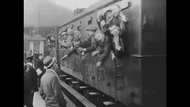 1910s: Train station.  Men lean out windows and wave.  Woman runs alongside train hold man's hand.  Soldiers march through town.  Family walks through field.