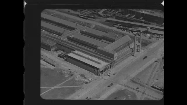 1920s: Looking down on factory buildings on large complex from sky above. Stream and smoke rise from factory buildings. Inside factory, car parts on assembly line.