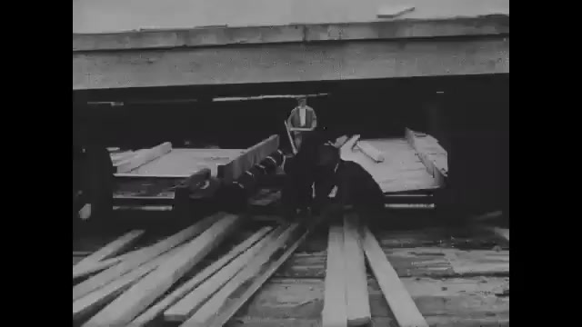 1910s: Men in saw mill pull lumber from conveyor belt. Intertitles. People work assembly line in automotive factory, car parts come down line. Man on assembly line. People working line.