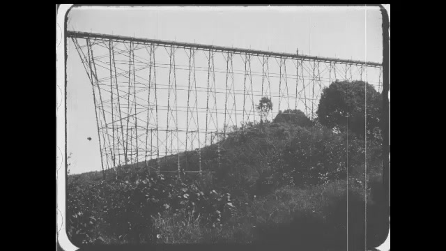 1910s: Person runs across water chute going over valley. Intertitles. Boat off shore approaches loading docks. Intertitles. Person walks holding bucket through forest.