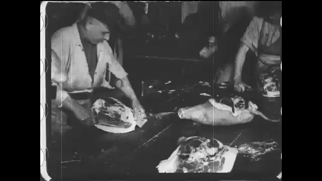 1910s: Workers trim fat from ham cuts. Intertitle. Worker uses a long handled cleaver to cut a shoulder from a pork side. 