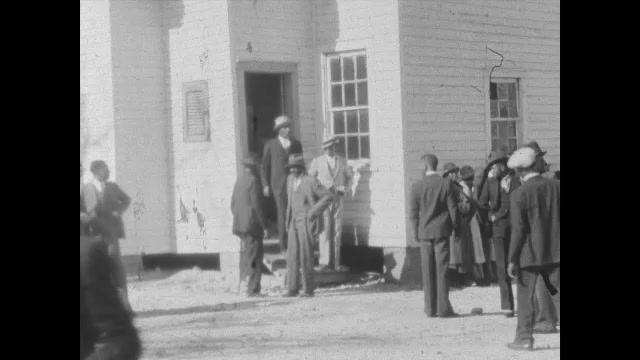 1930s: People stand in groups outside building. Couple stands outside house.