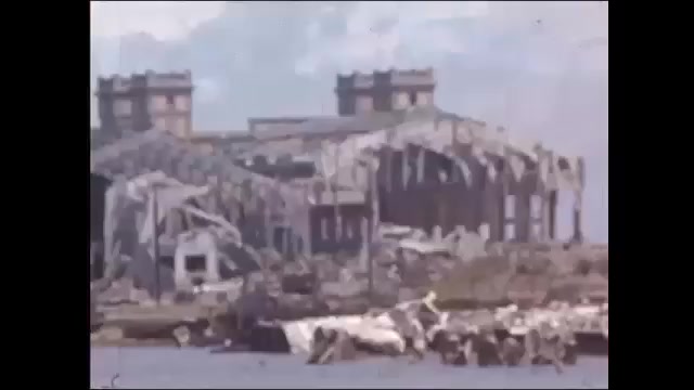 1940s: Destroyed shipping warehouse. Small fishing port. Soldier standing on damaged bridge. Woman with flowers, group waves. Long line of German military men walking along street.