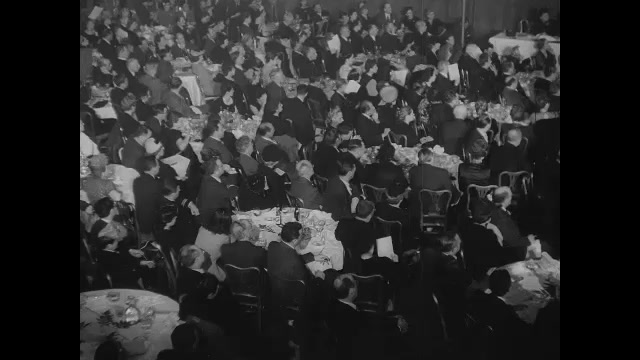 1940s A crowd of people sitting around tables listen to a speech. The crowd claps.