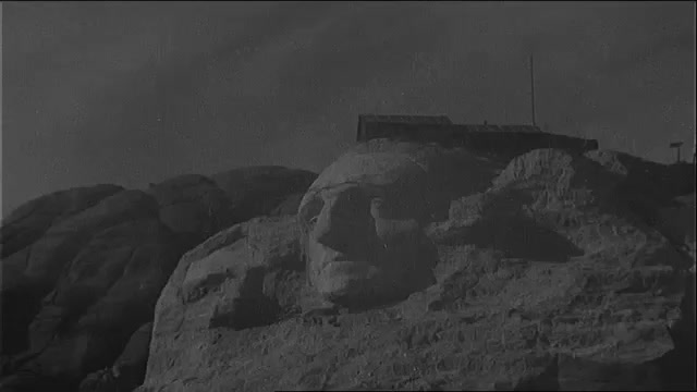 1930s: Blasting near George Washington head on Mount Rushmore. Workers climb near the head. Suspended workers carve the rock. Workers swing near the nose. People view the sculpture.