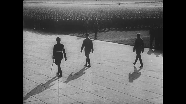 1930s: Adolph Hitler and two officers walk across stadium in Nazi rally. Crowd watches. Swastikas on flags, poles. Fire burns on pillars.