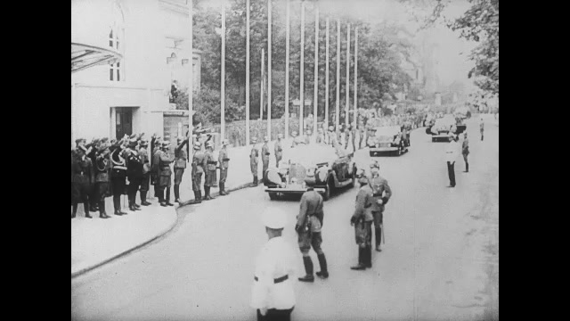 1930s Germany: Nazi officials exit car, enter building. Auditorium, man speaking. Officers in audience. Man speaking. Tracking shot, crowd waving to passing trucks. Crowd saluting soldiers in trucks. 