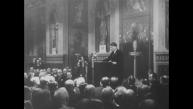 1930s Germany: Adolph Hitler stands behind podium and speaks to large audience in building.