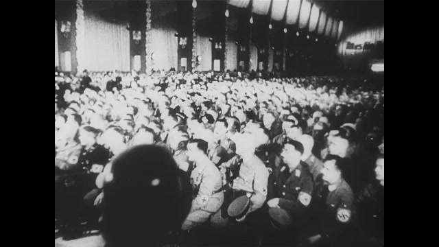 1930s Germany:  Crowd in hall with swastika symbols. Rudolf Hess in uniform speaks. Crowd of soldiers in uniform and people under Nazi Eagle symbols listen and rise to their feet. Adolf Hitler stands.