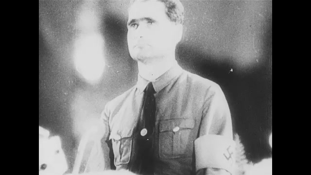 1930s Germany:  Huge crowd in hall with swastika symbols. Rudolf Hess in uniform speaks. Men listen, nod and clap. Man in uniform addresses the Fuhrer. 