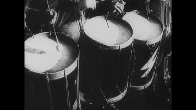 1930s Germany: Drum Corp beats their snare drums. Men in Nazi uniform hold shovels and chant. Adolf Hitler watches. Individual soldiers call out 