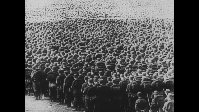 1930s Germany: Adolf Hitler stands on a platform above thousands of soldiers sitting in rows. Soldiers stand with shovels and flags, listening to Hitler speak. 