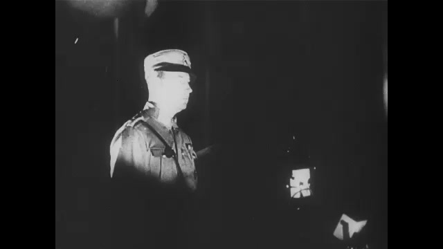 1930s Germany:  Officer in Nazi uniform speaks from dais.  Waving swastika flags. Nazi soldiers stand in the dark, burning torches and giving the Nazi salute. Close-up of a trumpet bell blowing. 