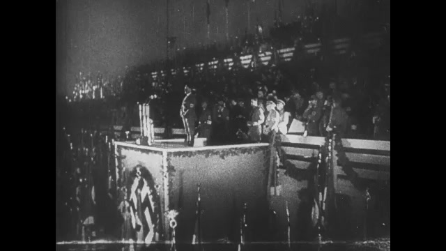 1930s Germany: Adolf Hitler stands on stage, gives speech. Soldiers in audience hold Nazi flags. 