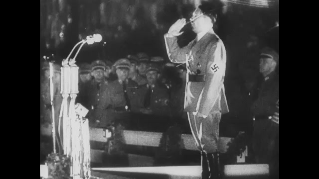 1930s Germany: Adolf Hitler stands on stage, gives speech, saultes. Crowd chants. Soldiers in audience hold Nazi flags. 