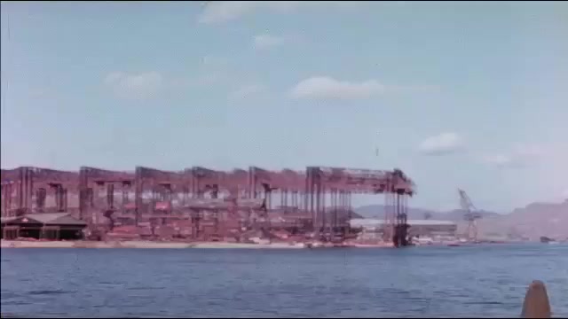 1940s: Empty shells of warehouses on shore. Small boat on ocean next to port. Row of empty cargo boats by shore.