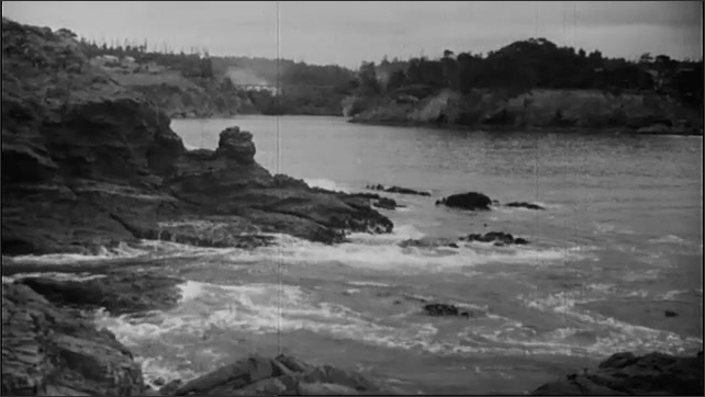1930s: Two cars drive on a dirt road overlooking an ocean inlet. Waves lap on a rocky shore. Men pick up debris around an inlet. Men pick up driftwood from a beach.
