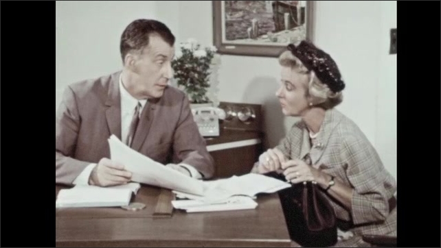1960s: Woman looks at papers and speaks. Man opens desk and pulls out papers. Man speaks.