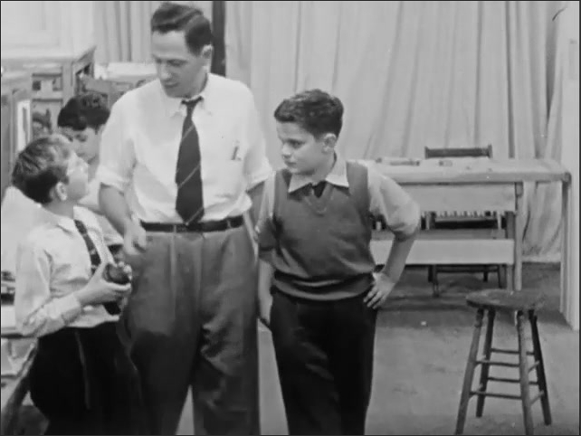 1950s: Man talking with boy in workshop, introducing boy to other children. Boys working with tools, materials in workshop. Boy taking off, hanging up jacket. 