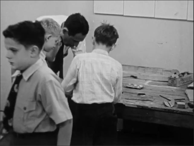 1950s: Man walking through workshop, holding piece of wood. Boys working at tables in workshop. Boys hammering objects at workbench. Man helping boys at work table.