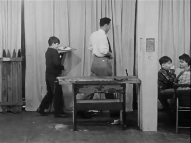 1950s: Boy walking around group of boys sitting at dinner table. Boy serving food to group. Man cleaning workbench. Man talking to boy, sitting down at dinner table.