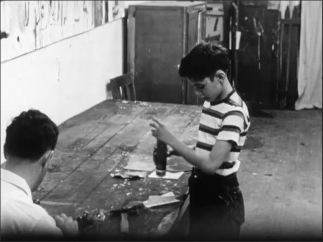 1950s: Boy talking to man sitting at work table. Boy removing rolled paper from tube, replacing paper, walking around workshop.
