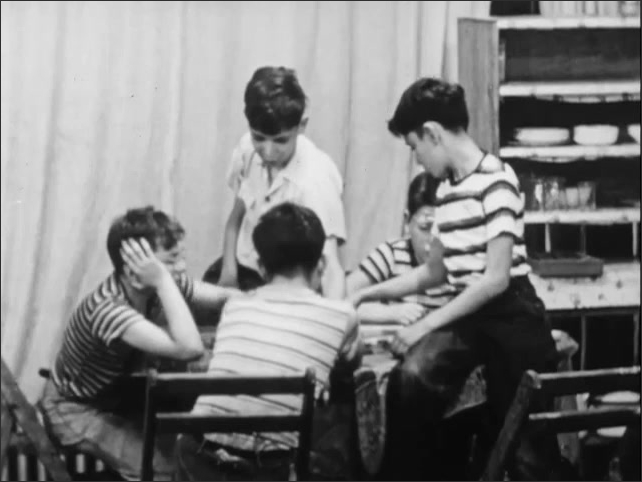 1950s: Boys playing checkers at table. Group of boys sitting at table, talking. Man holding dustpan talking to boys. Man sitting at table eating food with children, talking.
