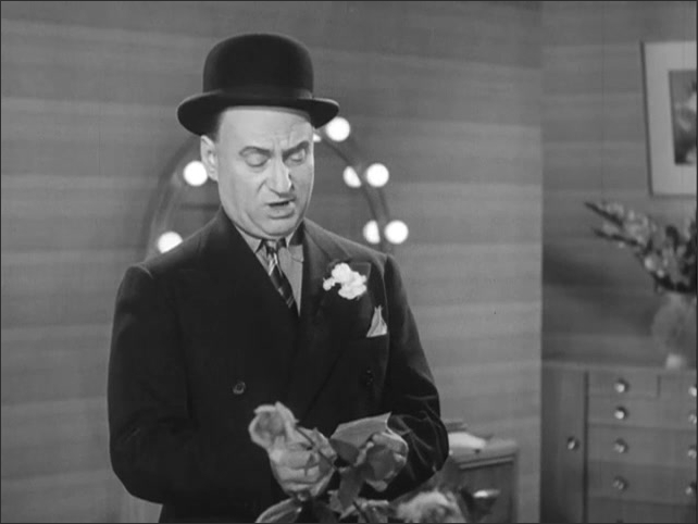 1940s: Woman holds bouquet of flowers, talks to man standing in doorway of dressing room. Man leaves. Woman puts flowers in vase while man talks standing next to her.