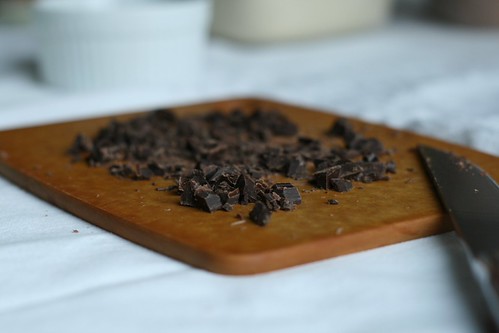 A bunch of chocolate chunks on a cutting board with a recently used knife to cut it.