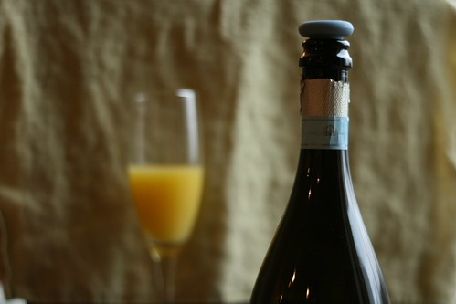 A stoppered bottle of champagne in the foreground with a background flute half full of orange juice.
