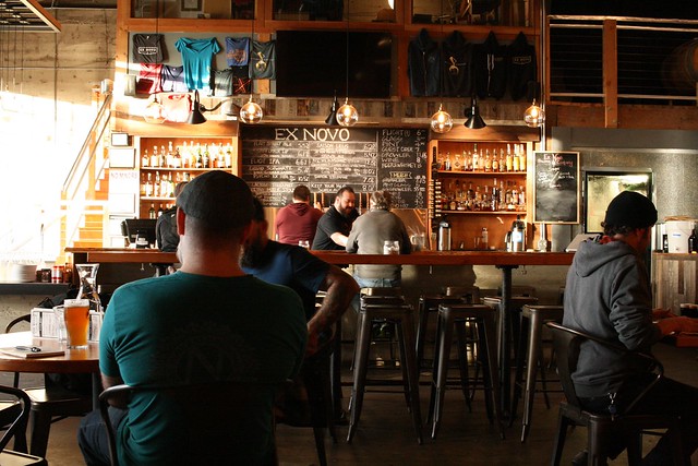 An indoor shot of Ex Novo brewing. You can see, but not read, the beer list in the far background, and there are two bartenders and four customers around.