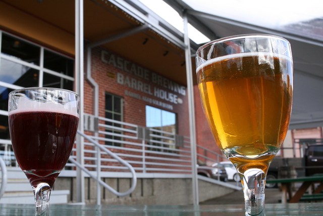 Two small goblets, one with golden beer and one with garnet beer on either side of the shot, with the name of the brewery, Cascade Brewing Barrel House, in the distance between them.