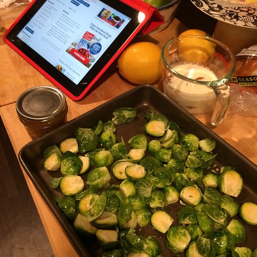 Foreground is a bunch of halved brussels sprouts in a sheet pan. Background is a measuring cup of cream, a canning jar of mustard, two lemons, and an iPad with a recipe up.