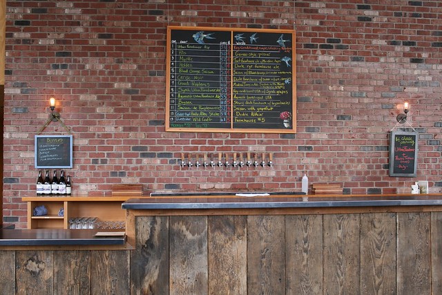 An empty bar, clean and very tidy. The taplist is centered over the taps which are, in turn, centered with regards to the bar. On either side there's a chalkboard listing something.