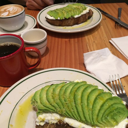 A table with avocado toast nearest the camera and another avocado toast in the background. A cup of coffee is near the nearer toast and a cappuccino is near the further toast. 