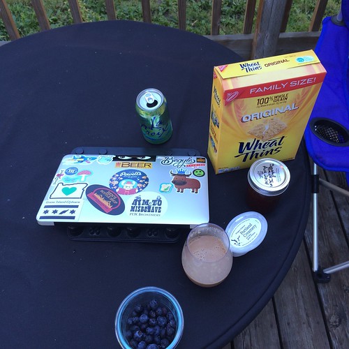 A closed laptop , covered in stickers, on a table next to a chocolate milk, some berries, some jam, and some wheat thins crackers.